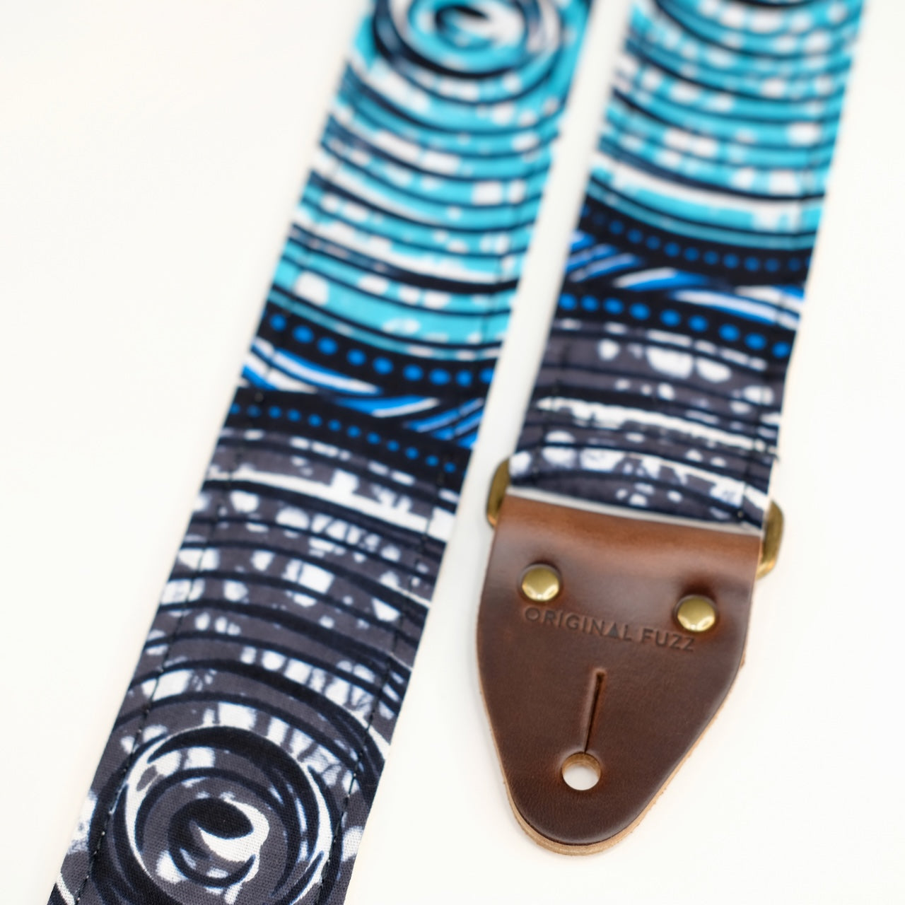 End-tab detail view of guitar strap in white, black, blue, teal, and gray swirled African wax print fabric with brown leather end-tab. Made in Nashville by Original Fuzz.