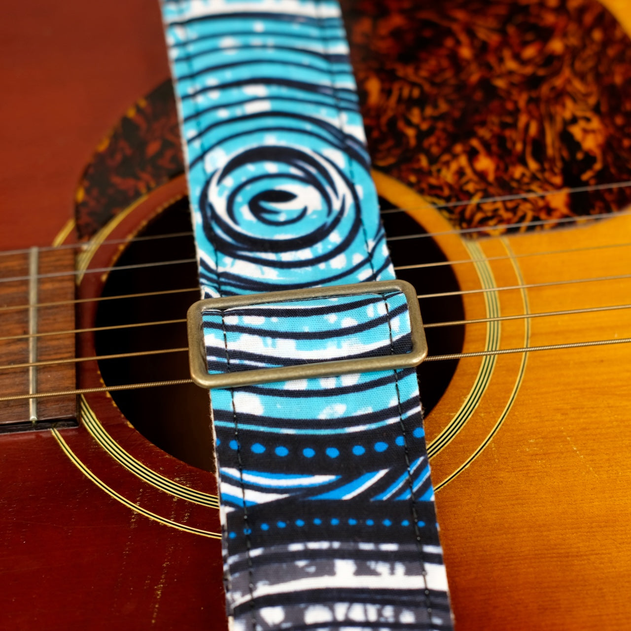 Buckle detail of guitar strap in white, black, blue, teal, and gray swirled African wax print fabric with brown leather end-tab. Made in Nashville by Original Fuzz.