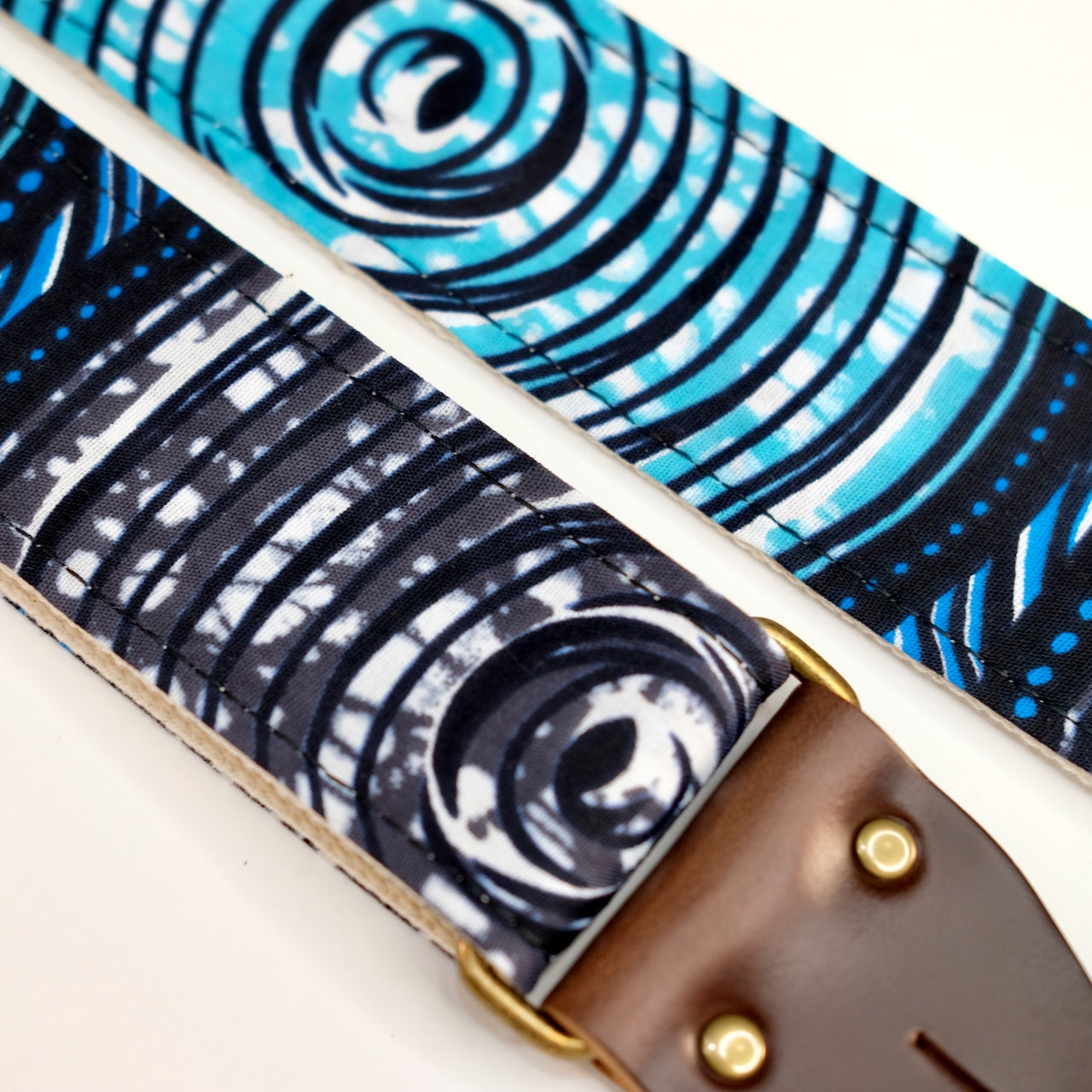 Fabric detail view of guitar strap in white, black, blue, teal, and gray swirled African wax print fabric with brown leather end-tab. Made in Nashville by Original Fuzz.