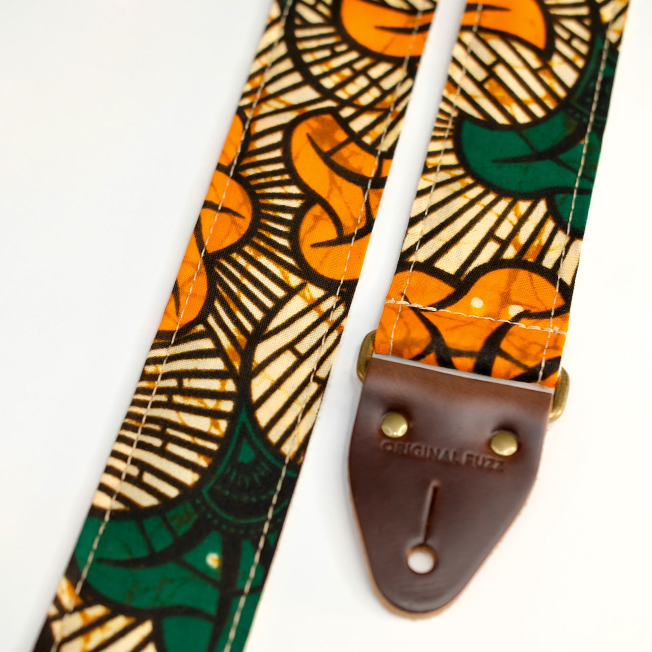 End-tab detail view of guitar strap in tan, orange, and green botanical African wax print fabric with brown leather end-tab. Made in Nashville by Original Fuzz.