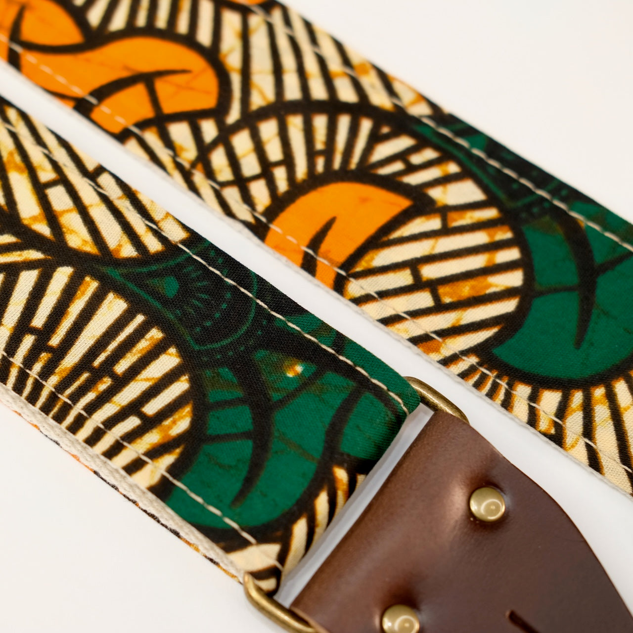 Fabric detail view of guitar strap in tan, orange, and green botanical African wax print fabric with brown leather end-tab. Made in Nashville by Original Fuzz.
