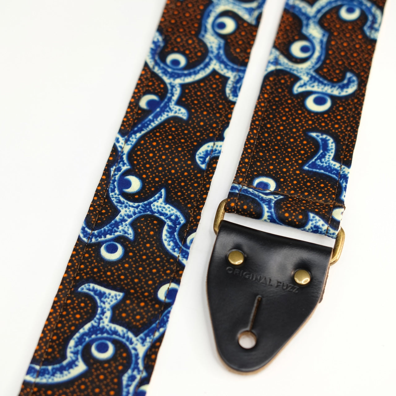 End-tab detail view of guitar strap in black, blue, cream, and orange botanical African wax print fabric with black leather end-tab. Made in Nashville by Original Fuzz.