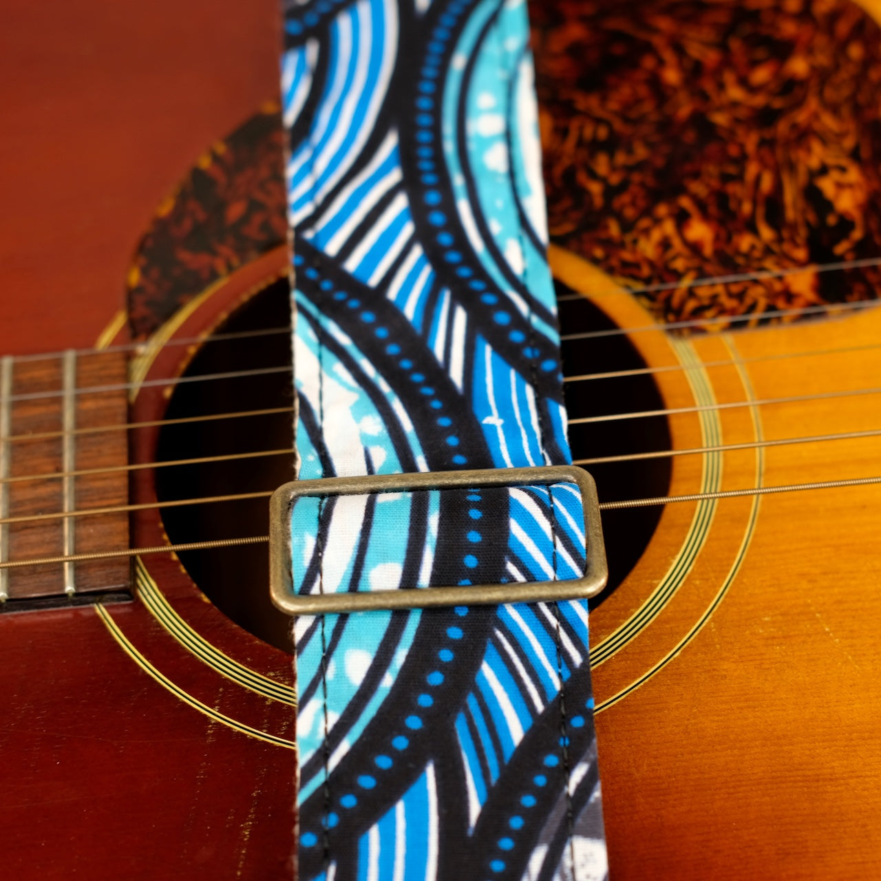 Buckle detail of guitar strap in teal, gray, white, black, and blue rippled African wax print fabric with brown leather end-tab. Made in Nashville by Original Fuzz.