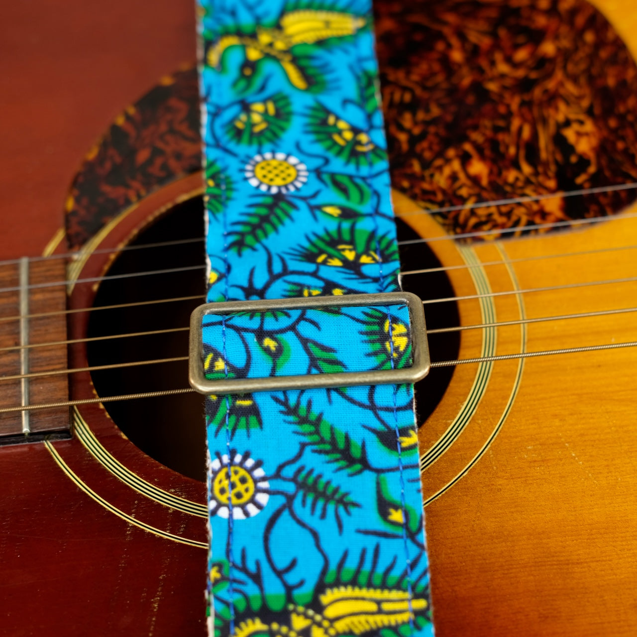 Buckle detail of guitar strap in blue, yellow, and green botanical African wax print fabric with brown leather end-tab. Made in Nashville by Original Fuzz.
