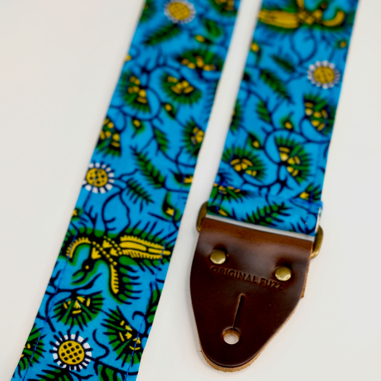 End-tab detail view of guitar strap in blue, yellow, and green botanical African wax print fabric with brown leather end-tab. Made in Nashville by Original Fuzz.