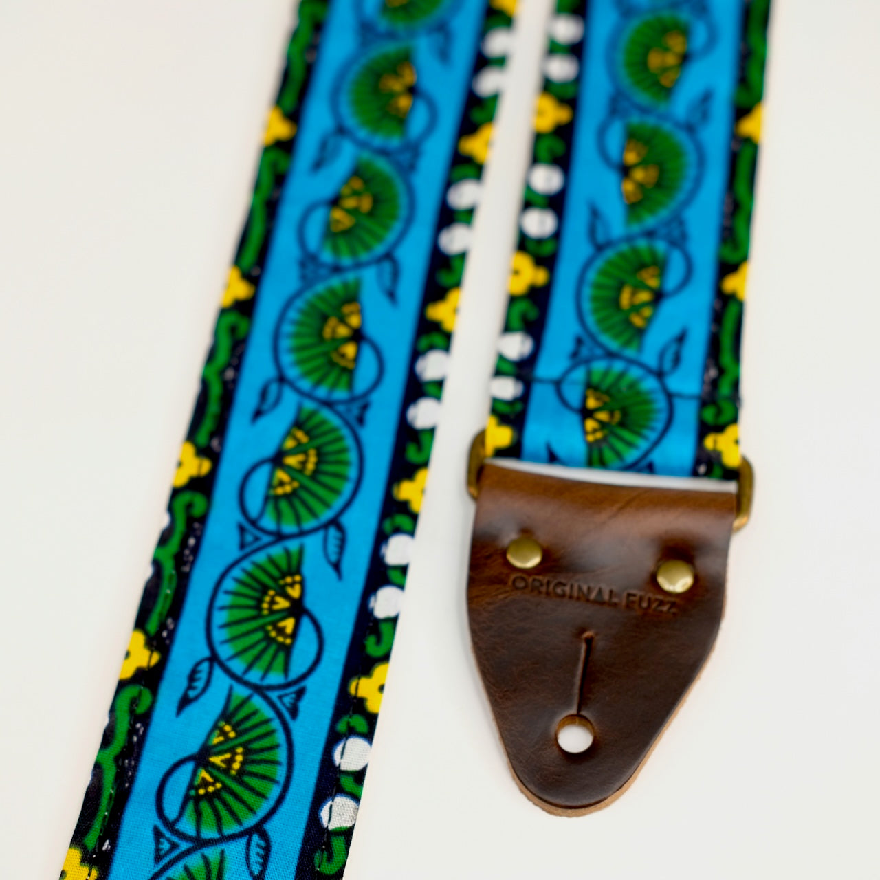 End-tab detail view of guitar strap in blue, yellow, and green bordered botanical African wax print fabric with brown leather end-tab. Made in Nashville by Original Fuzz.