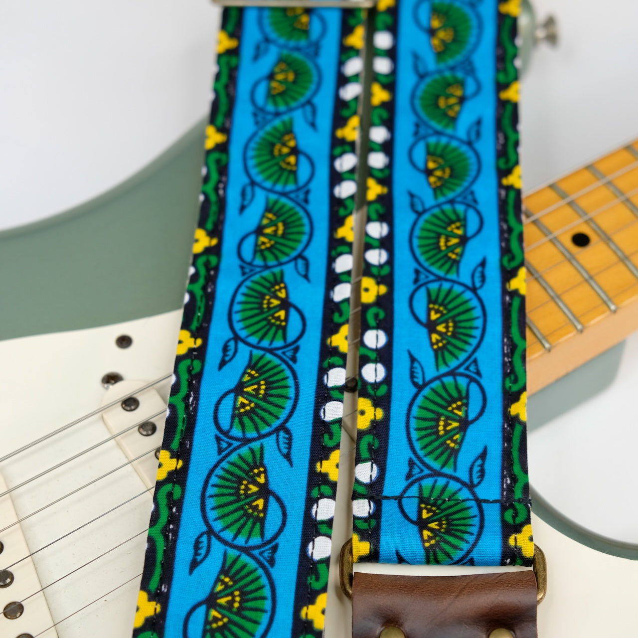Wide view of guitar strap in blue, yellow, and green bordered botanical African wax print fabric with brown leather end-tab. Made in Nashville by Original Fuzz.
