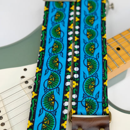 Wide view of guitar strap in blue, yellow, and green bordered botanical African wax print fabric with brown leather end-tab. Made in Nashville by Original Fuzz.